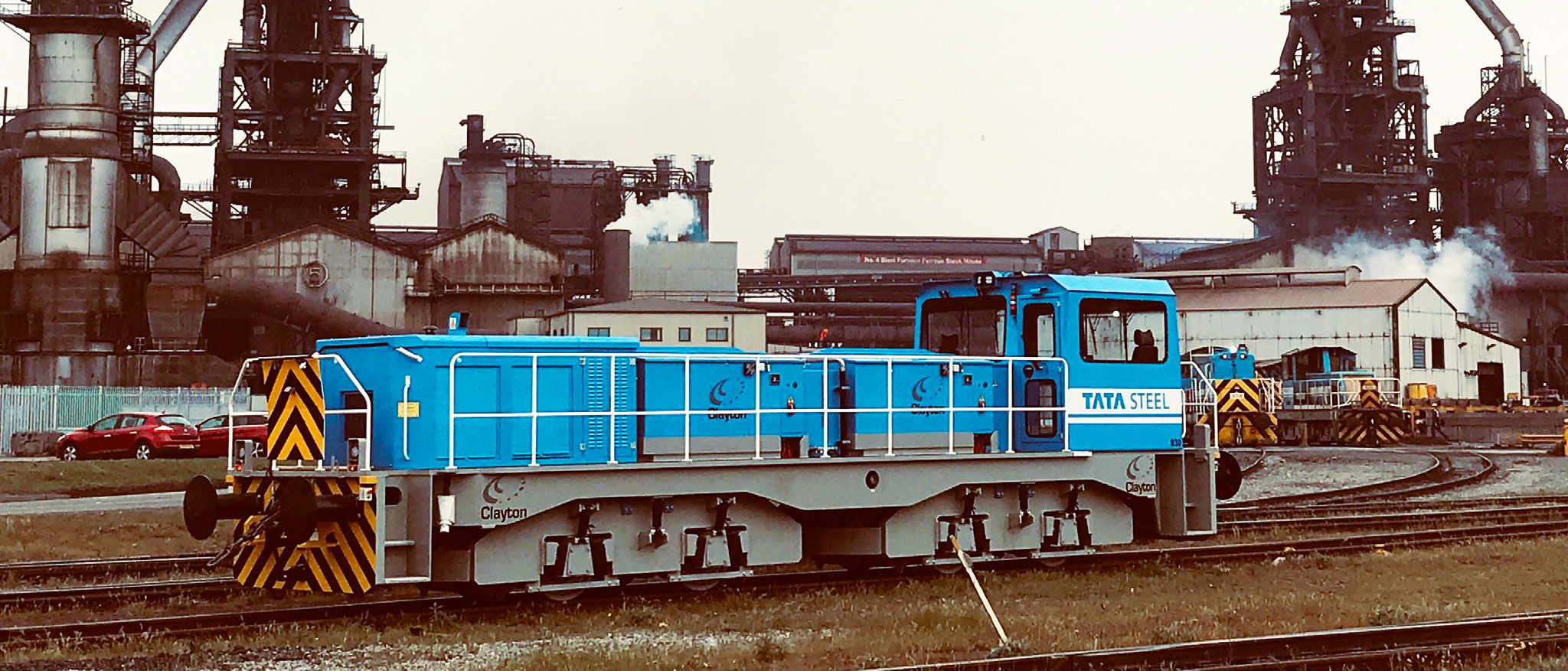 A blue hybrid locomotive driving past an industrial area.